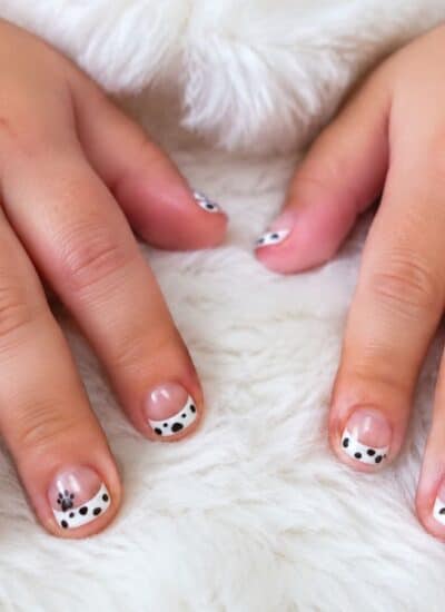 A pair of hands with short nails painted with black and white Dalmatian dog designs resting on a white furry surface.
