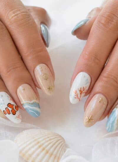 Hands with long nails featuring ocean-themed nail art, including waves, coral, a clownfish, seashells, and blue and beige colors. Seashells and a starfish are in the background.