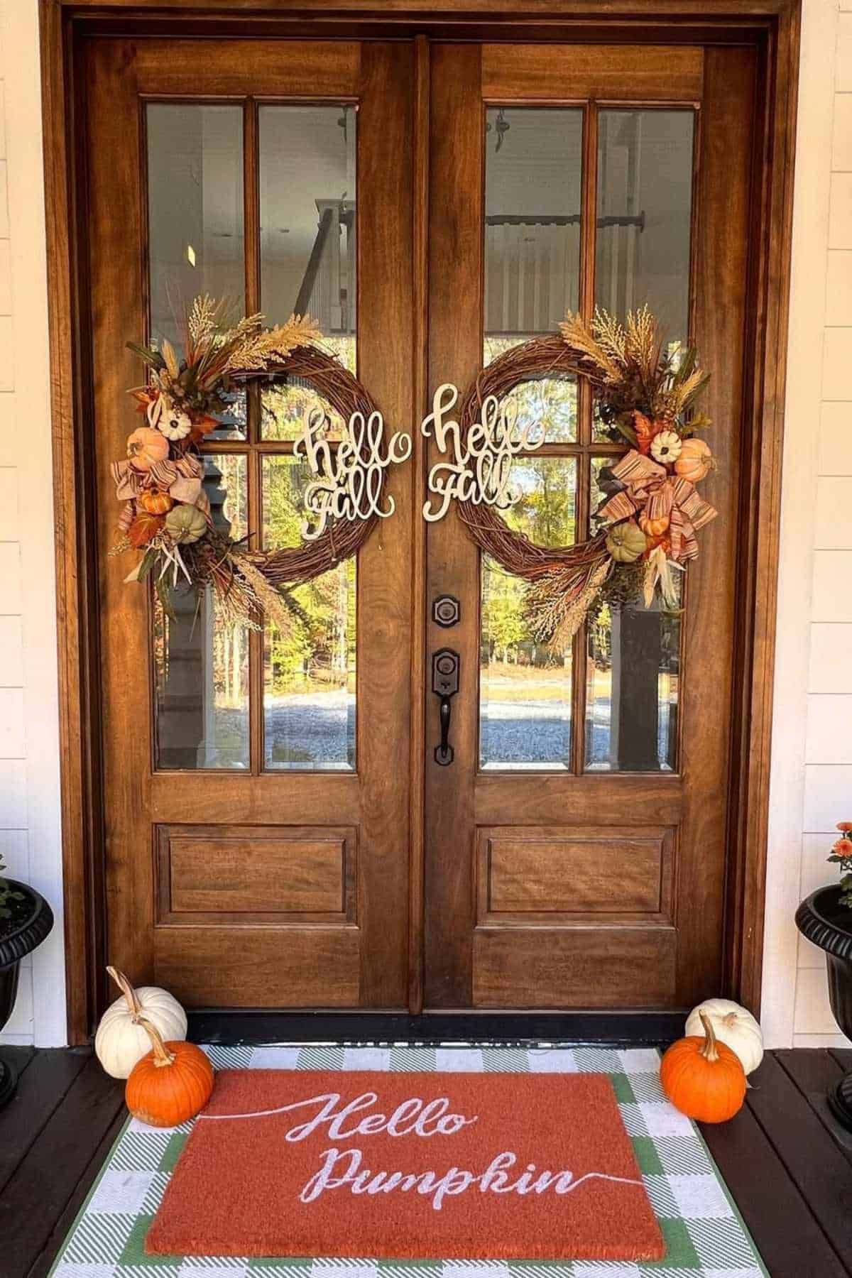 Double wooden doors decorated with fall wreaths and "hello fall" signs; autumn-themed doormat reads "Hello Pumpkin" with pumpkins arranged on either side.