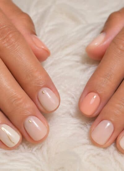 Two hands with short, neatly manicured nails in a pale pink and peach gradient, each nail featuring a small gold accent, resting on a white textured surface.