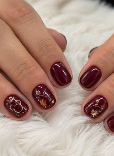 Hands with short, oval dark red nails featuring gold and white leaf and abstract designs, resting on a white textured surface.