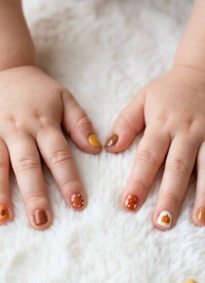 A child’s hands with autumn-themed nail art rest on a white surface, surrounded by small pumpkins, acorns, and fall leaves.