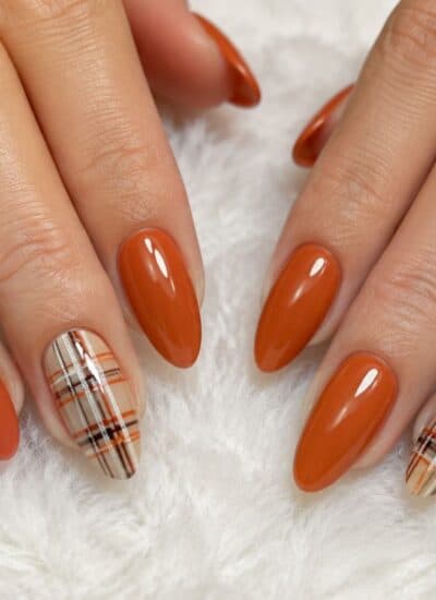 Hands with almond-shaped nails painted orange, with two accent nails on each hand featuring an orange, black, and white plaid pattern, resting on a white fluffy surface.