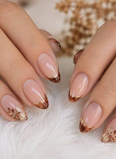 Hands with manicured nails featuring brown French tips, delicate gold floral designs, and gold rings on a soft white surface.