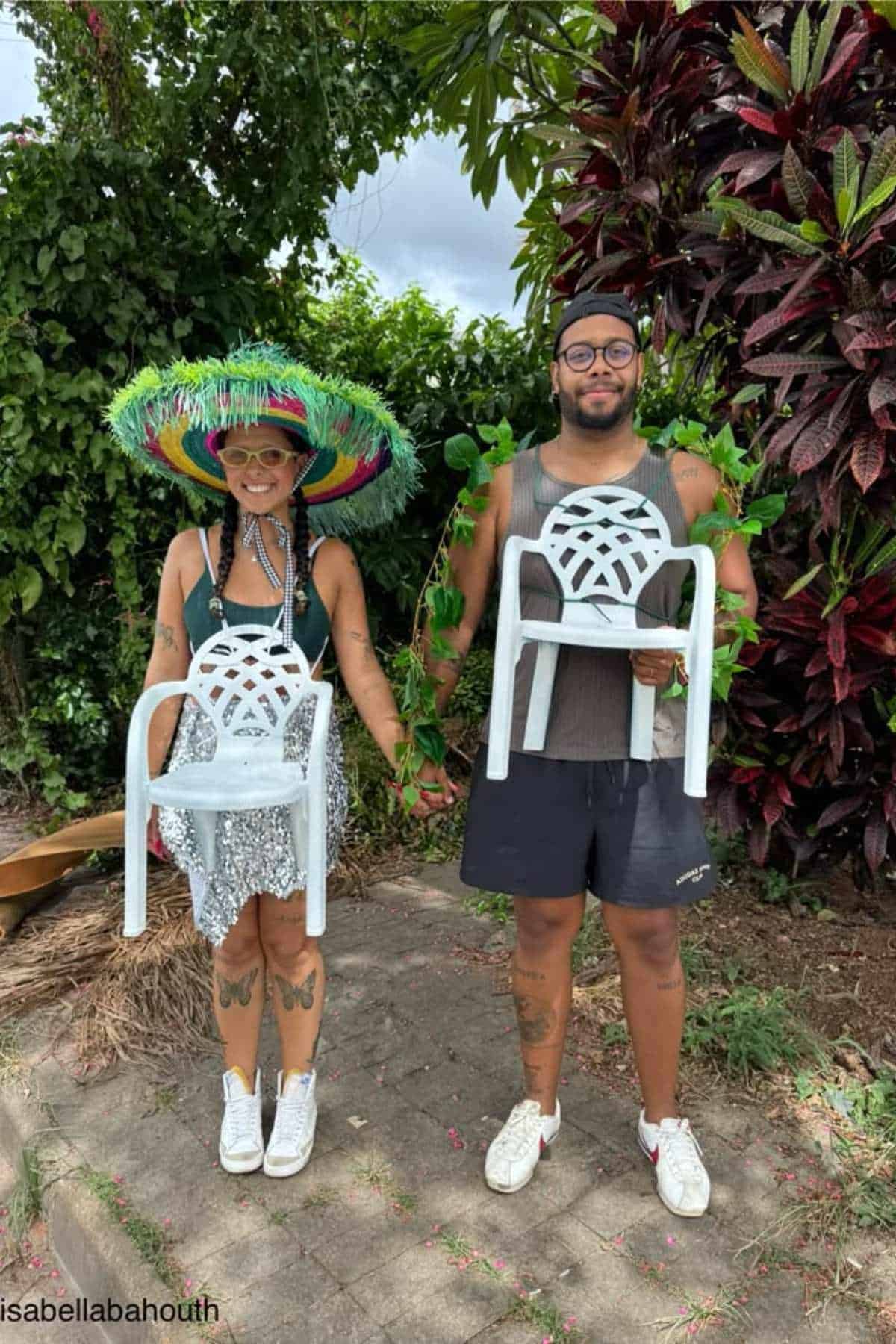Two people stand outdoors holding small white plastic chairs. One person wears a large colorful hat, and both have greenery draped around their arms. They are smiling and holding hands.