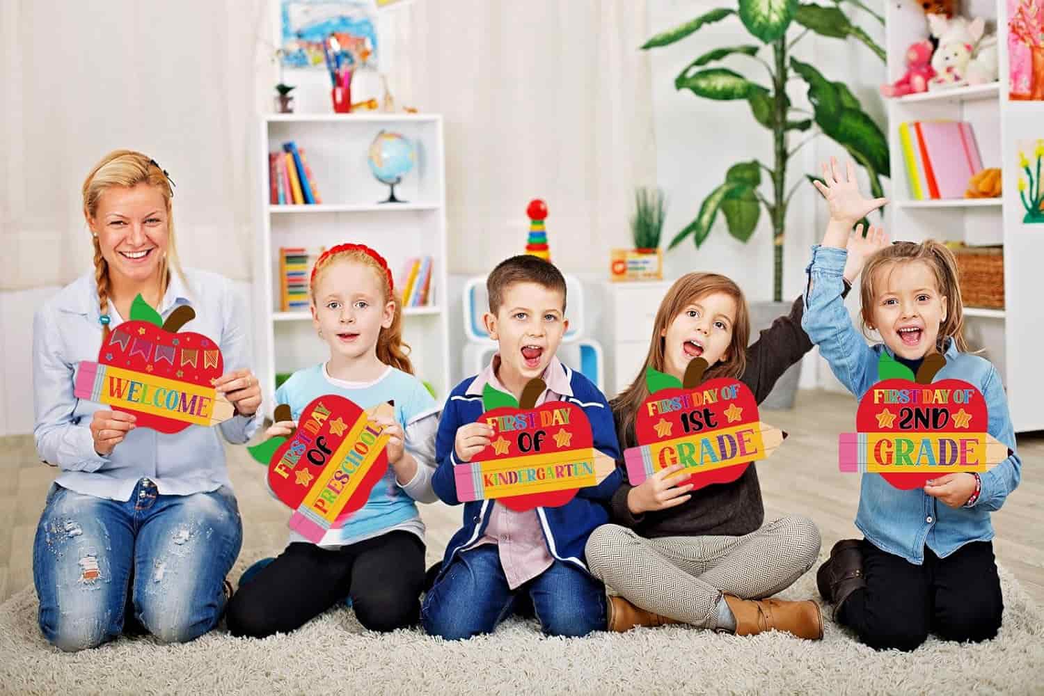A teacher and five young children sitting on the floor, holding colorful signs that mark the first day of various school grades in a classroom setting.