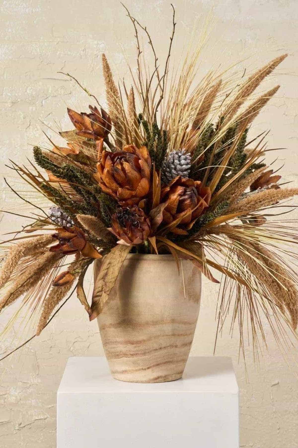 A beige ceramic vase holds a dried flower arrangement with brown, orange, and tan flowers, grasses, and seed pods against a neutral background.