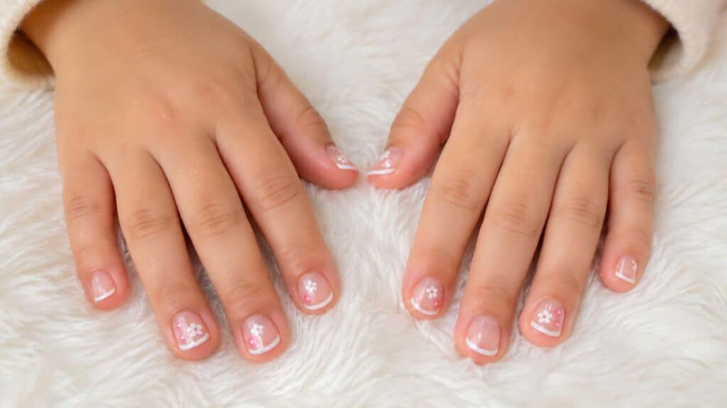 Two hands with manicured nails featuring a simple French tip design and small white flower decorations, resting on a white fluffy surface.