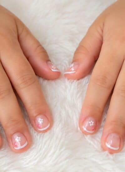 Two hands with manicured nails featuring a simple French tip design and small white flower decorations, resting on a white fluffy surface.