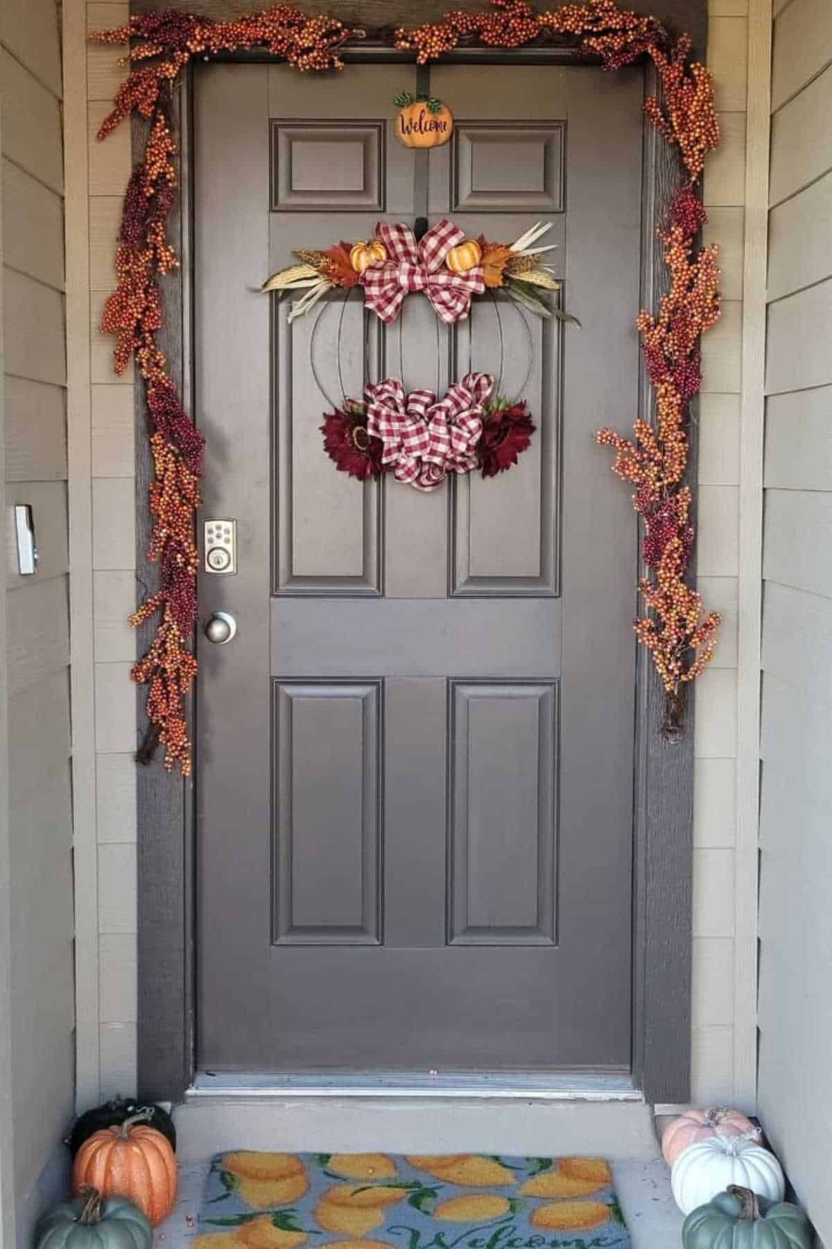 A gray front door decorated with a fall wreath, orange berry garland, and small pumpkins on the doorstep, with a welcome mat at the entrance.