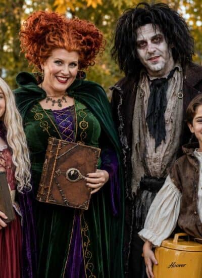 Four people in Halloween costumes pose outdoors; one dressed as a witch with red hair, one as a ghostly figure, and two children in Hocus Pocus-style outfits, one holding a vacuum.