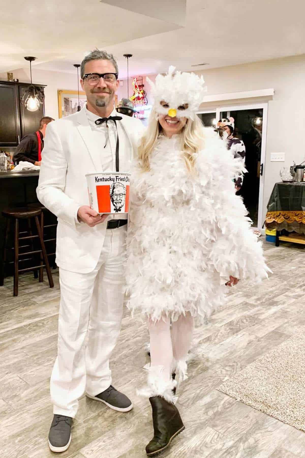Two people in a kitchen; one is dressed in a white suit holding a KFC bucket, and the other is wearing a fluffy white chicken costume.