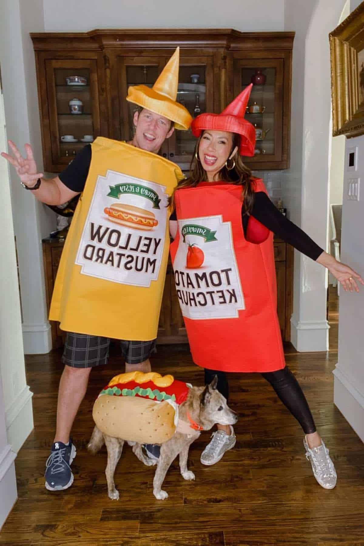 Two people wearing mustard and ketchup bottle costumes stand next to a dog dressed as a hot dog in a home setting.