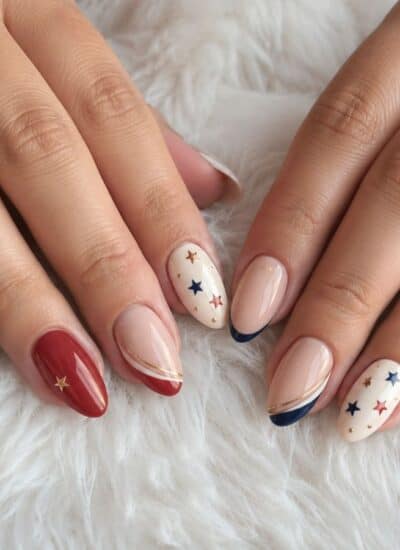 Two hands with manicured nails painted in red, navy blue, and beige, featuring star designs and accent lines, resting on a white, fluffy surface.