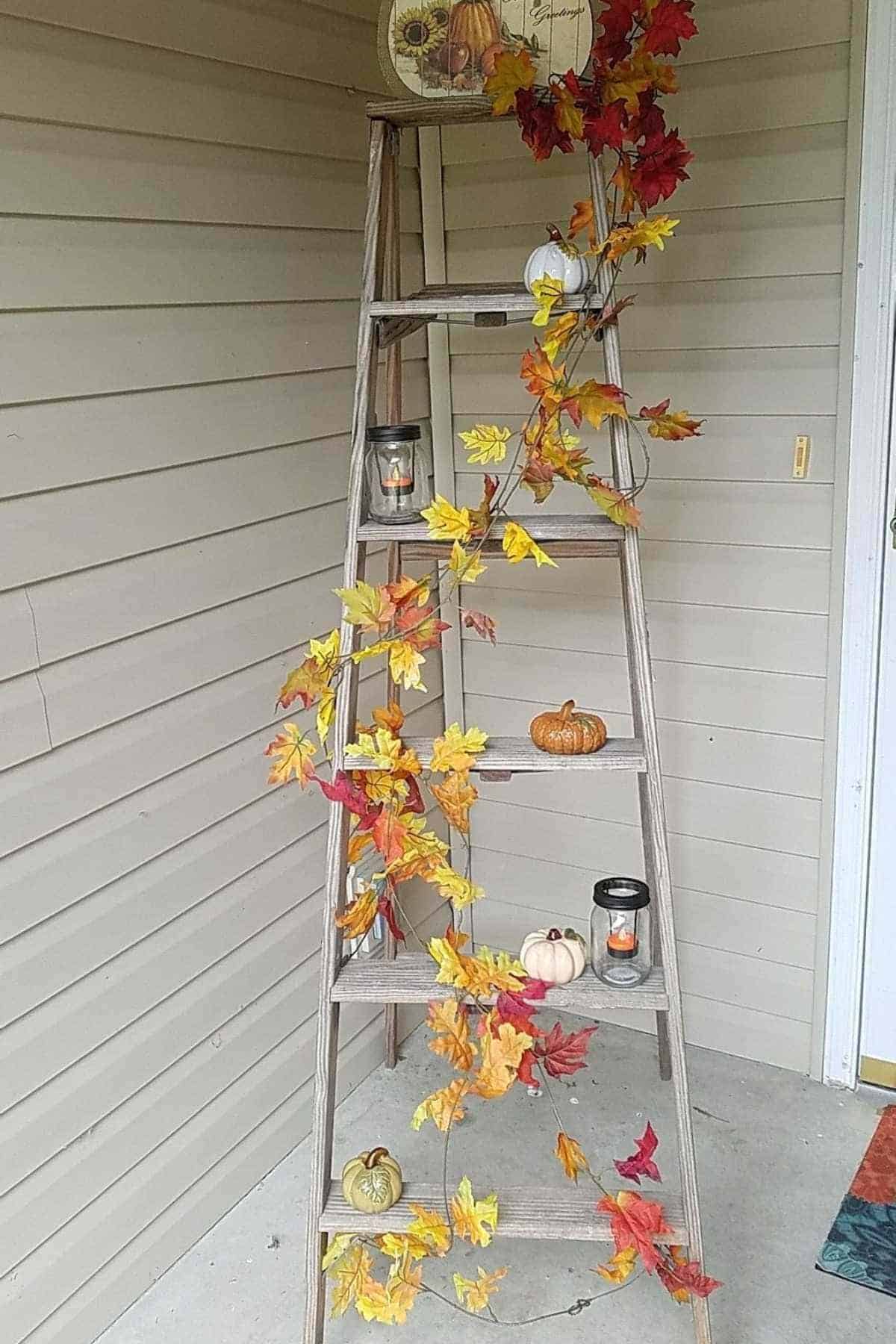 A wooden ladder decorated with artificial autumn leaves, small pumpkins, two lanterns with candles, and an autumn-themed plate on the top shelf, set against a beige siding wall.