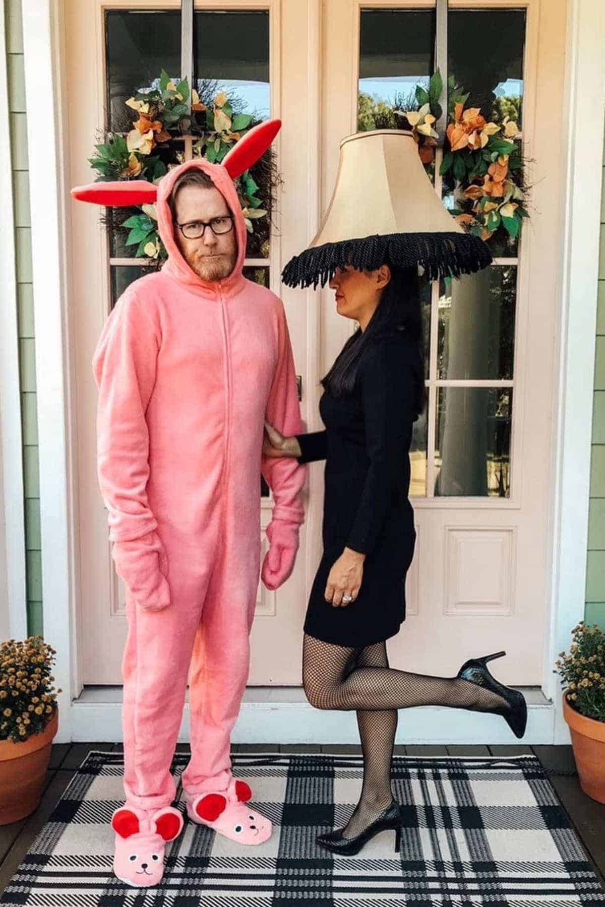 A man in a pink bunny costume stands next to a woman dressed as a lamp with a lampshade on her head, posing in front of double doors decorated with wreaths.