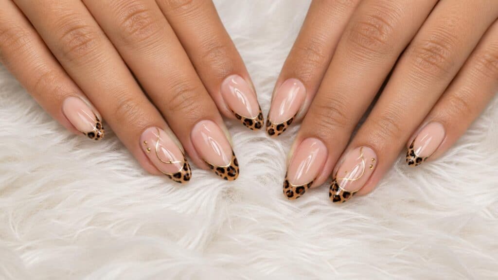 Close-up of hands with manicured nails featuring nude polish and leopard print French tips, accented with gold lines and small gold studs, resting on a white furry surface.