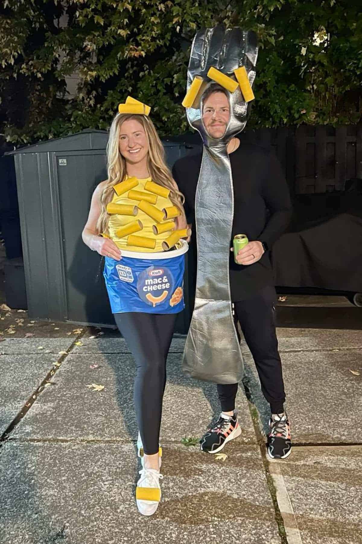 Two people in costumes stand outdoors; one is dressed as a box of mac and cheese, the other is dressed as a large fork with pieces of macaroni on it.