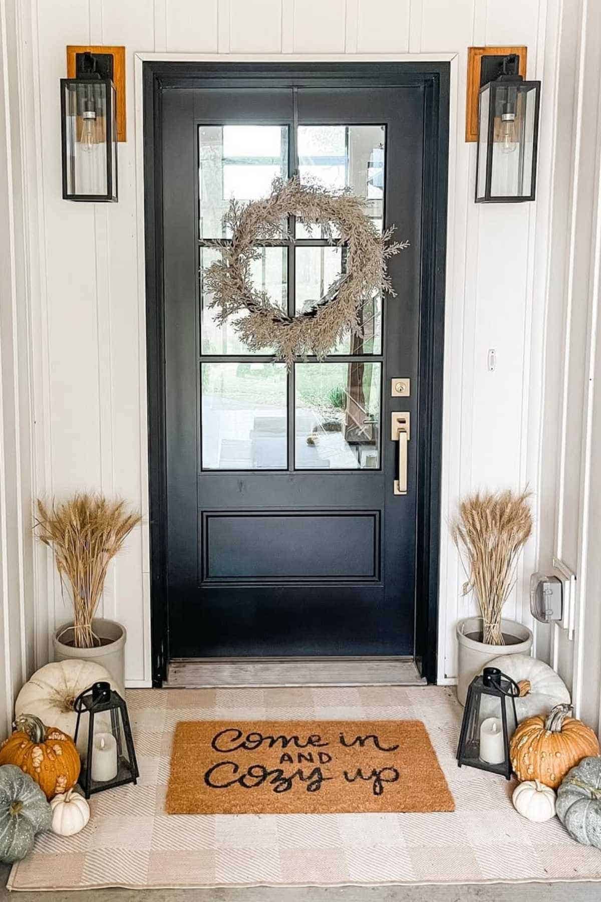 A black front door with a wreath, flanked by lanterns, wheat bundles, and pumpkins. A doormat reads, "Come in and cozy up.