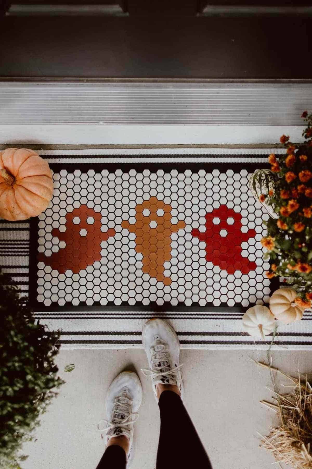 A person stands on a porch mat decorated with three cartoon ghosts; pumpkins and potted flowers are placed on either side of the mat.