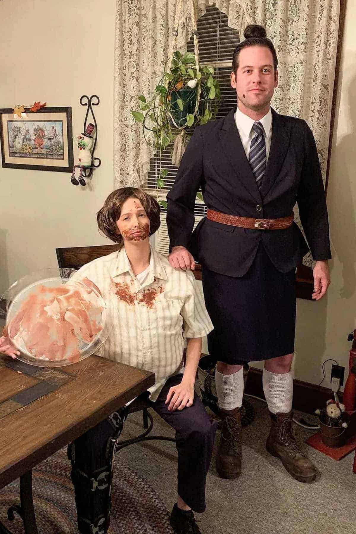 Two people in costume; one seated with a fake beard, dirty shirt, and plate of ham, the other standing in a suit jacket, skirt, boots, and knee socks. Indoors with houseplants and decor.
