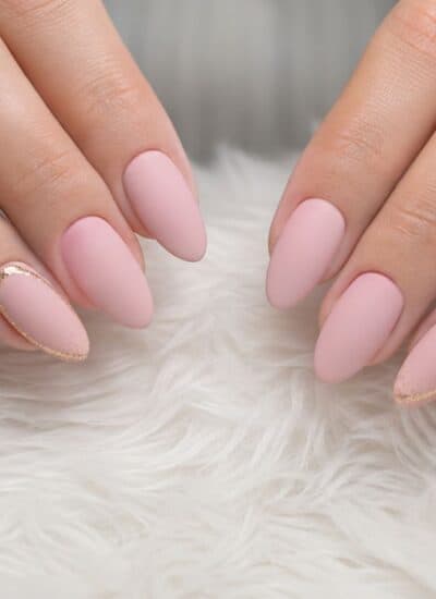 Hands with almond-shaped nails painted light pink, featuring gold glitter accents near the cuticles, resting on a white furry surface.