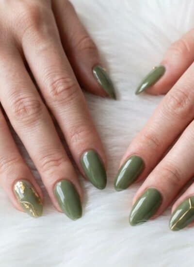 Two hands with almond-shaped nails painted olive green, featuring gold accents on the ring fingers, resting on a white, fluffy surface.