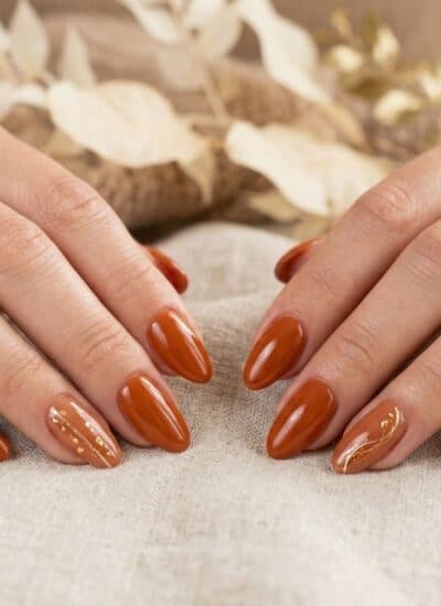 Two hands with almond-shaped nails painted in glossy brown polish, some decorated with gold accents, resting on a beige fabric surface with dried leaves in the background.