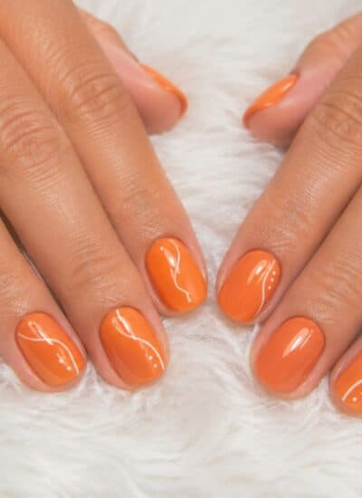 Two hands with neatly manicured nails painted in glossy orange polish rest on a white, fuzzy surface.