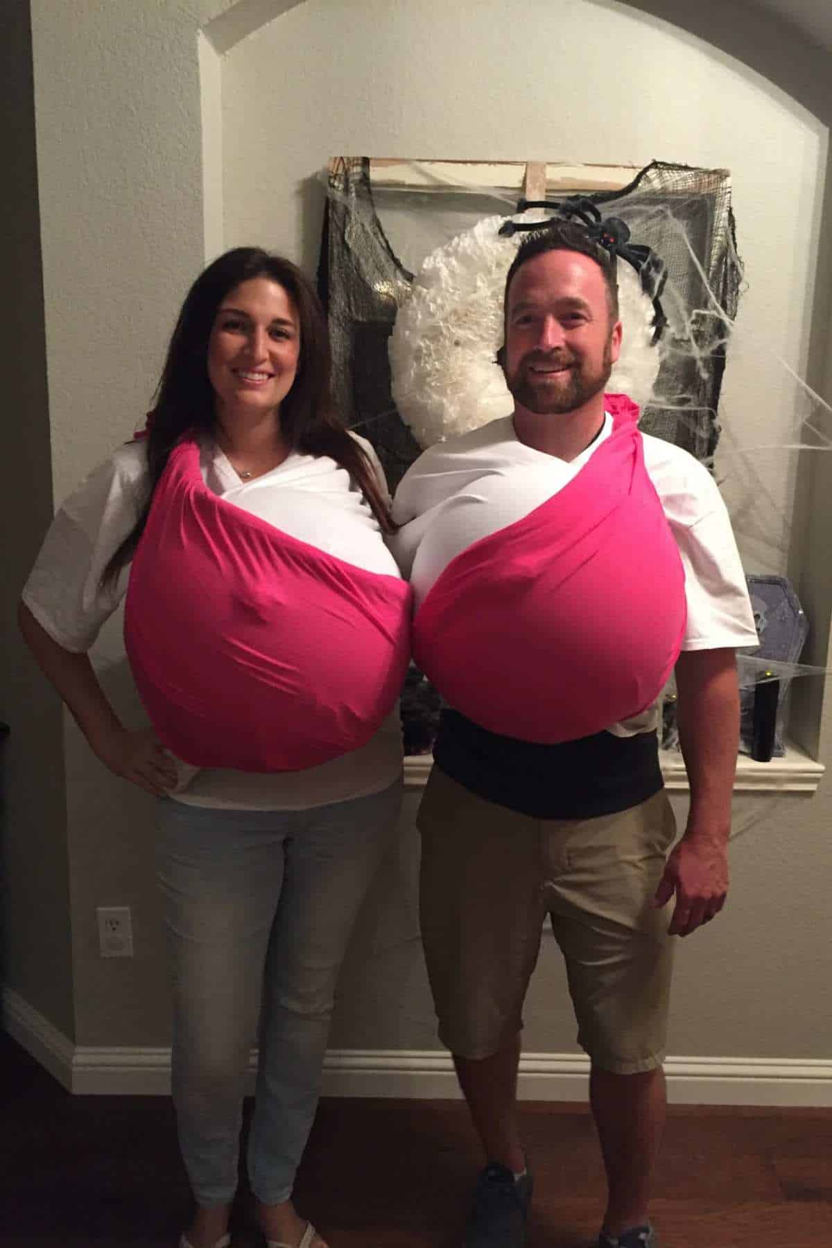 Two adults stand side by side, each wearing a large, pink and white costume that resembles exaggerated breasts. They are smiling and posing indoors.