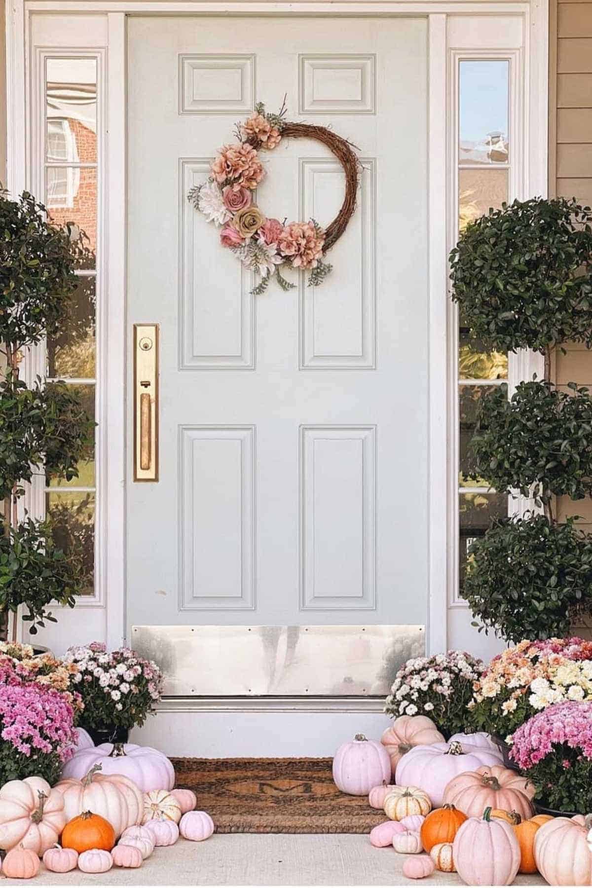 A white front door with a floral wreath, flanked by two potted shrubs. Pink and white pumpkins and potted flowers decorate the doorstep.