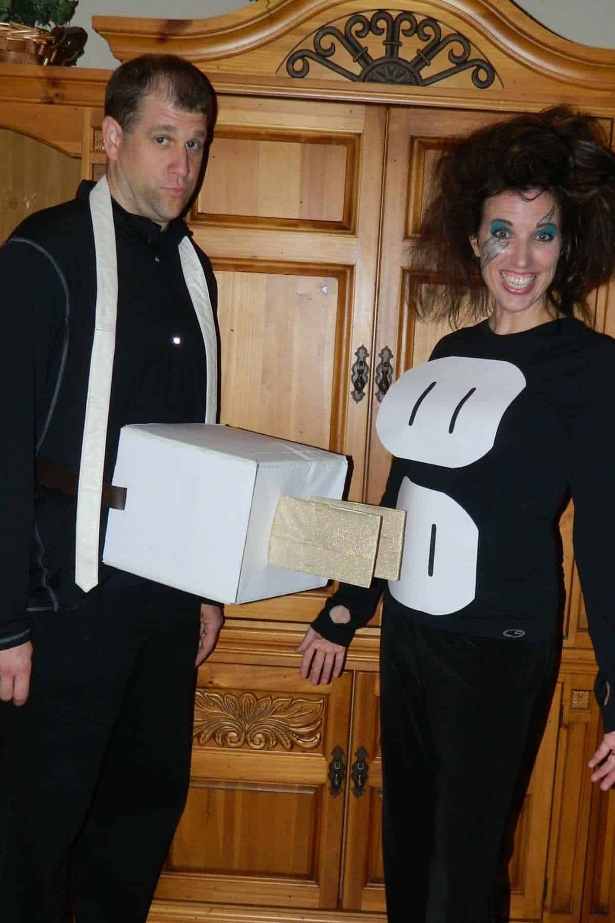 Two people in costumes stand indoors; one is dressed as an electrical plug, the other as an electrical outlet. Both are posing and smiling for the camera.