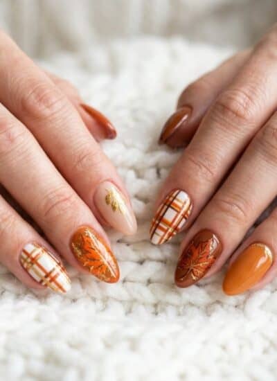 Hands with manicured nails painted in fall-themed colors and designs, including plaid patterns and leaf motifs, resting on a white knitted fabric.