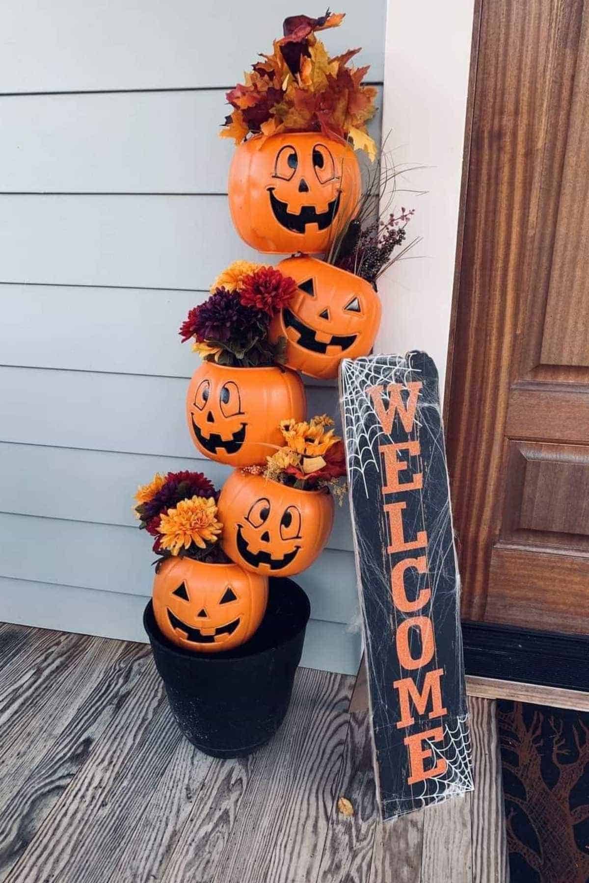 Stacked plastic jack-o'-lanterns with artificial flowers in a black pot next to a vertical "Welcome" sign decorated with spiderwebs by a front door.