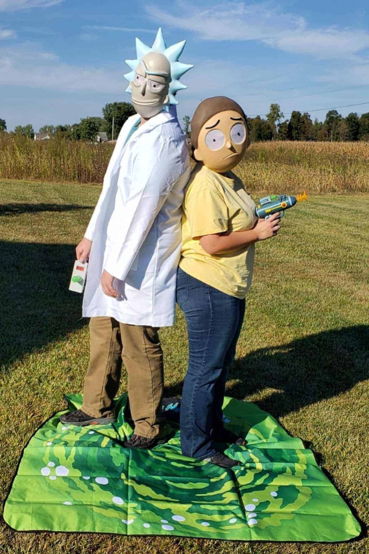 Two people in a grassy field wear Rick and Morty character costumes, standing back to back on a green portal-themed mat.