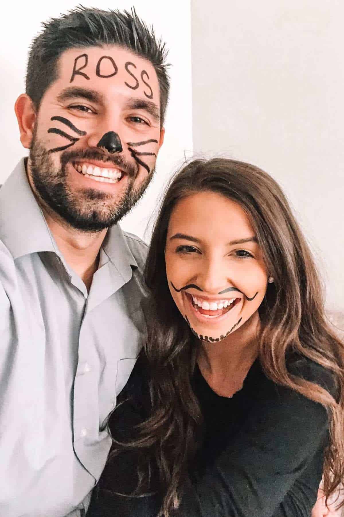 A man and woman smile at the camera with faces drawn on in marker; the man has "ROSS" on his forehead and whiskers, the woman has whiskers and "CHANDLER" on her chin.