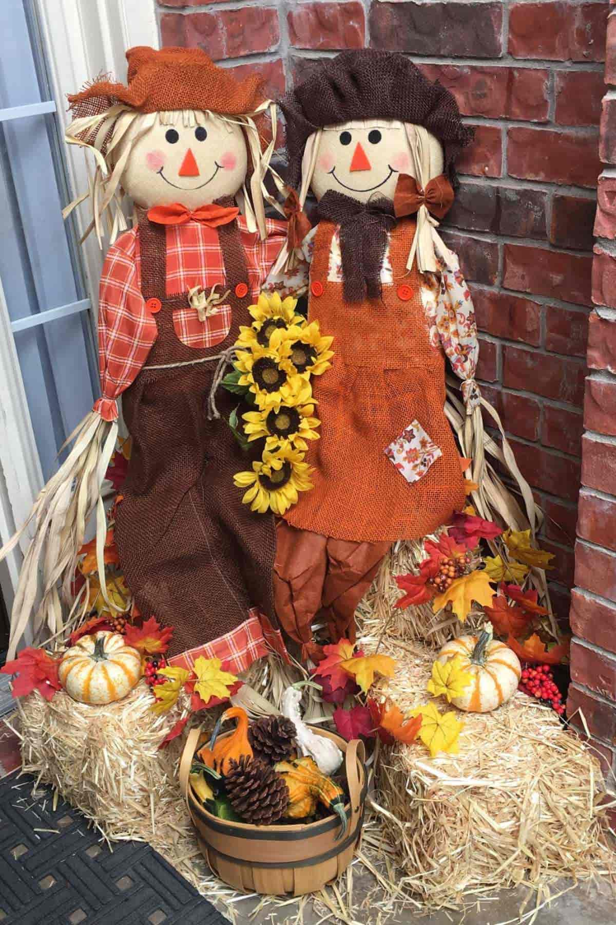 Two scarecrow dolls sit on hay bales surrounded by small pumpkins, artificial sunflowers, autumn leaves, and a basket with pinecones, set against a brick wall.