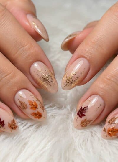 Hands with almond-shaped nails featuring nude polish, gold glitter tips, and autumn leaf designs resting on a white, fluffy surface.