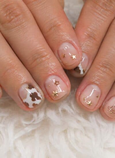 Hands with short nails featuring a clear base, brown and white cow print accents, and small gold star-shaped nail art, resting on a fluffy white surface.