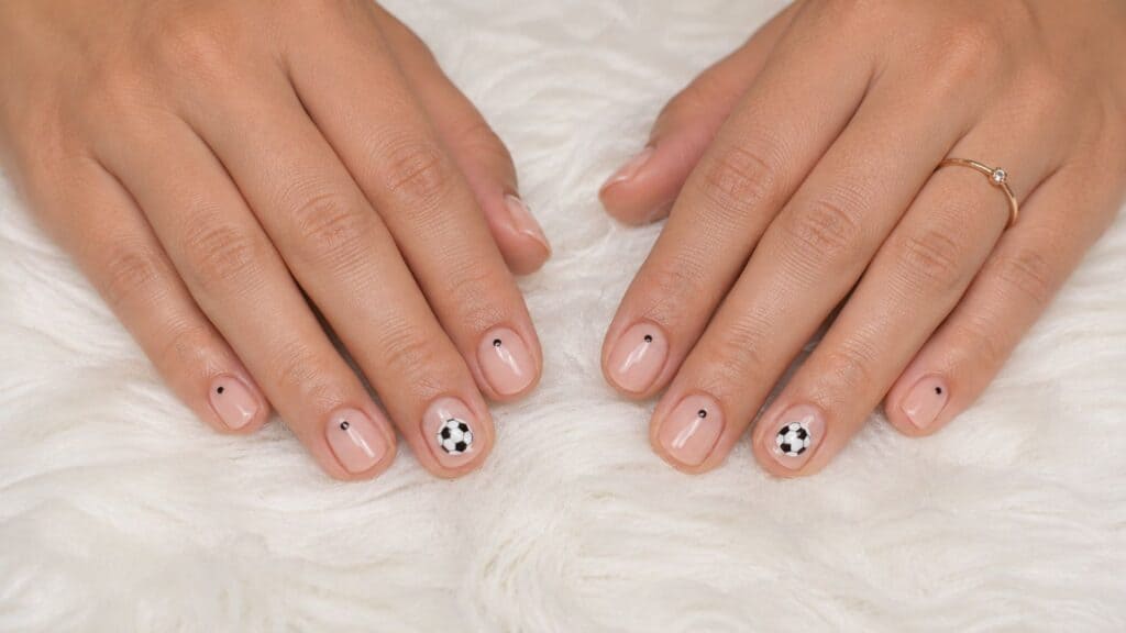 Two hands with nude nail polish, each nail featuring a small black dot; two nails have black and white soccer ball designs. Hands rest on a white, fluffy surface.