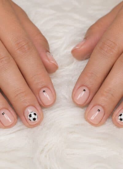 Two hands with nude nail polish, each nail featuring a small black dot; two nails have black and white soccer ball designs. Hands rest on a white, fluffy surface.