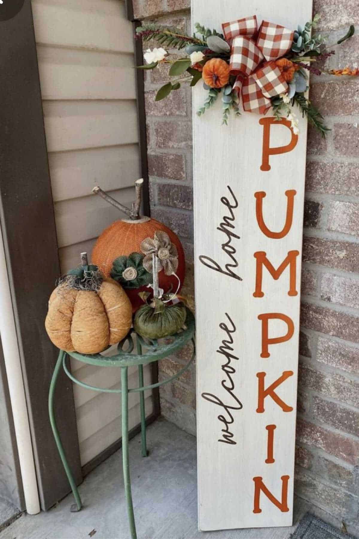 A decorative sign reading "welcome home PUMPKIN" stands next to a small table with pumpkins and fall-themed ornaments on a porch.