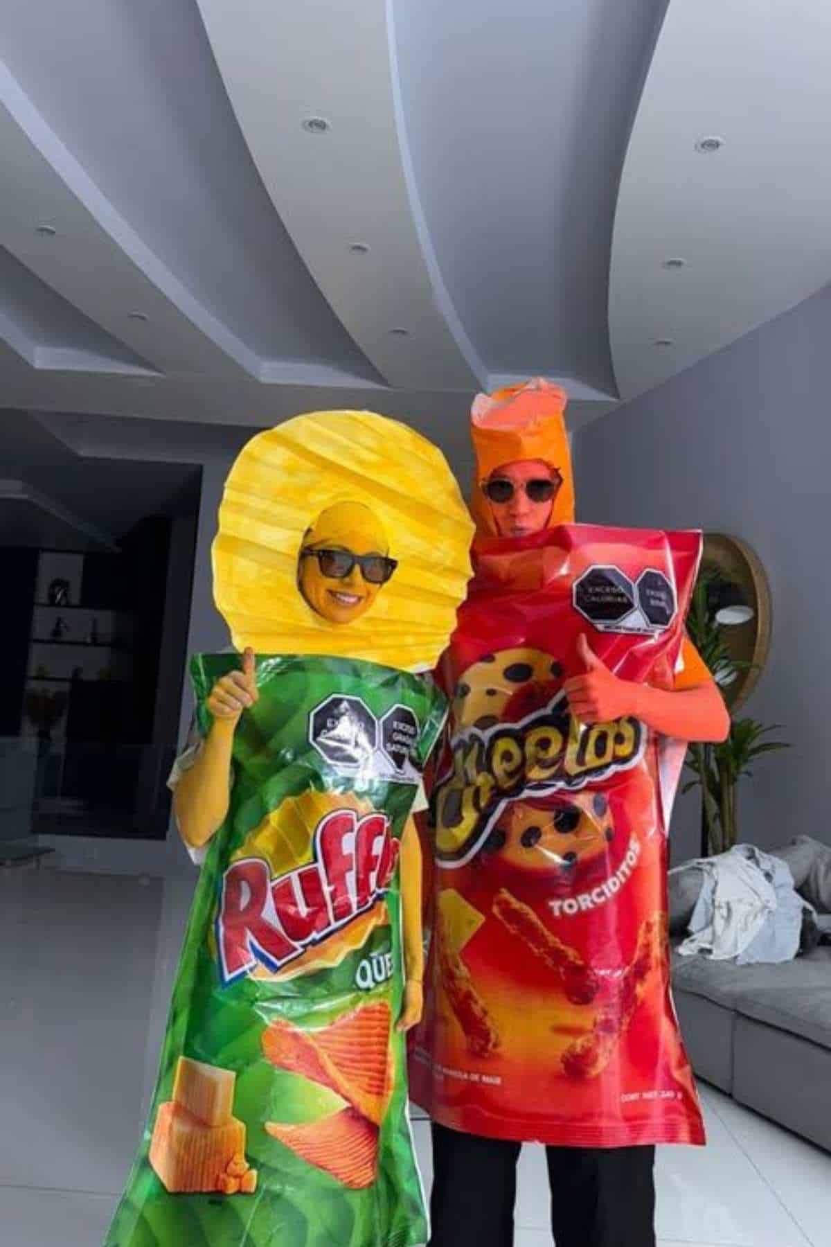 Two people wearing costumes resembling Ruffles and Cheetos snack bags stand indoors, both giving thumbs up and wearing sunglasses.