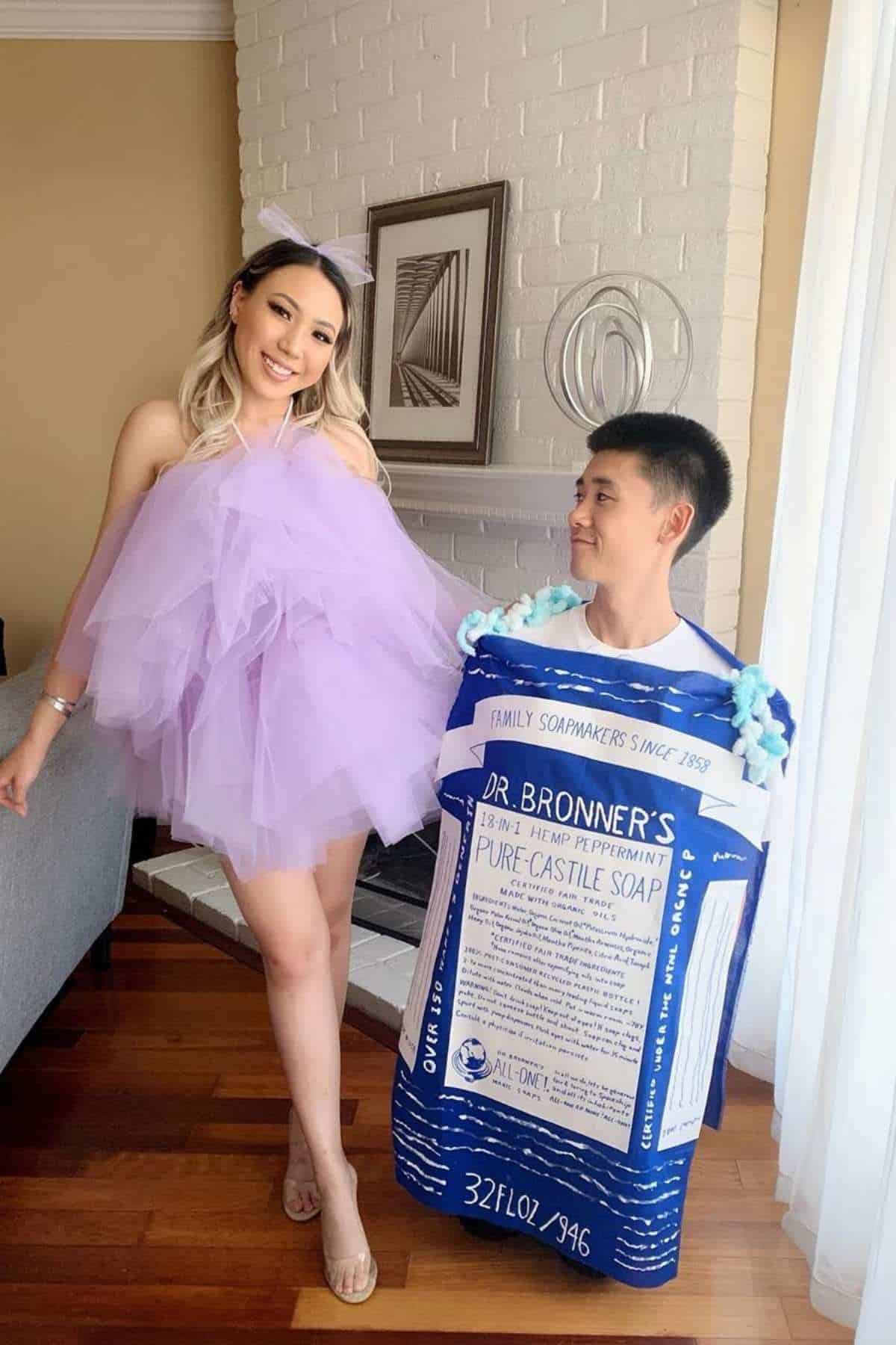 A woman in a purple tulle dress and a man in a Dr. Bronner’s soap bottle costume stand together indoors, smiling.