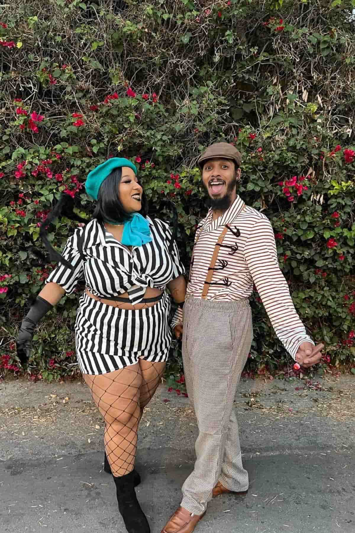 Two people in vintage-inspired, black-and-white striped costumes stand smiling in front of a leafy, flower-covered wall.