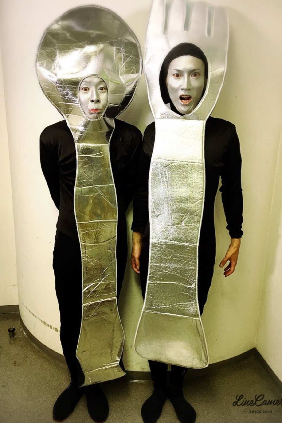 Two people wearing metallic spoon and fork costumes stand side by side against a white wall, with their faces visible through cutouts.