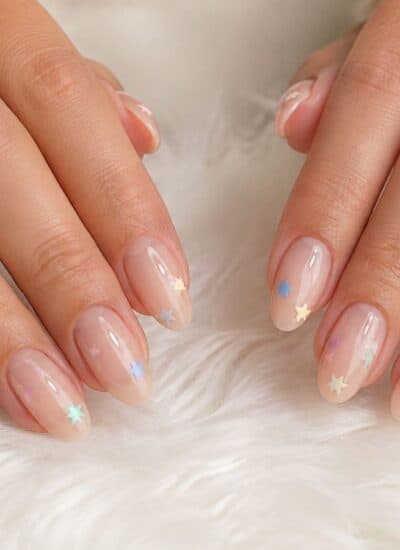 Two hands with manicured nails featuring a nude base and small, colorful star decals rest on a white, fluffy surface.