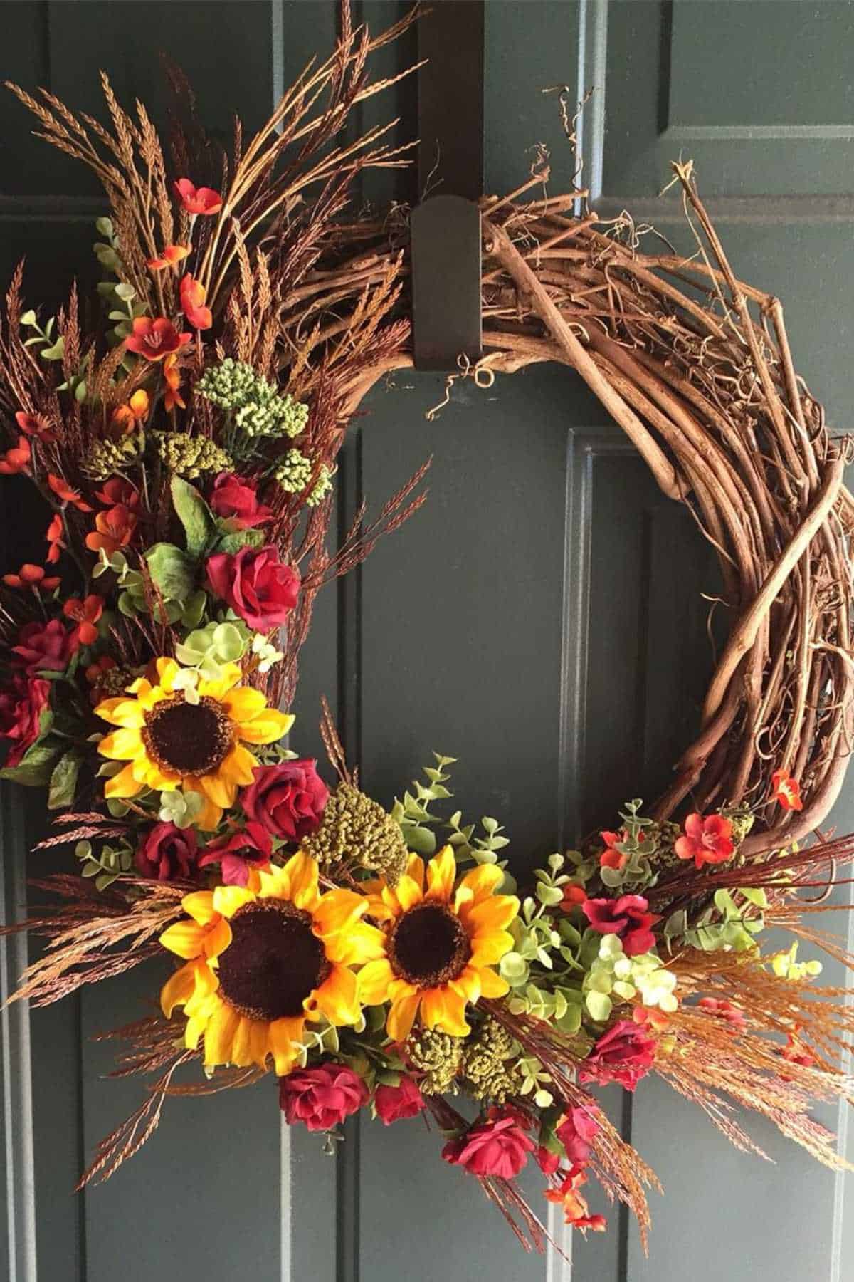 A grapevine wreath decorated with artificial sunflowers, red roses, greenery, and dried grasses hangs on a dark-colored door.