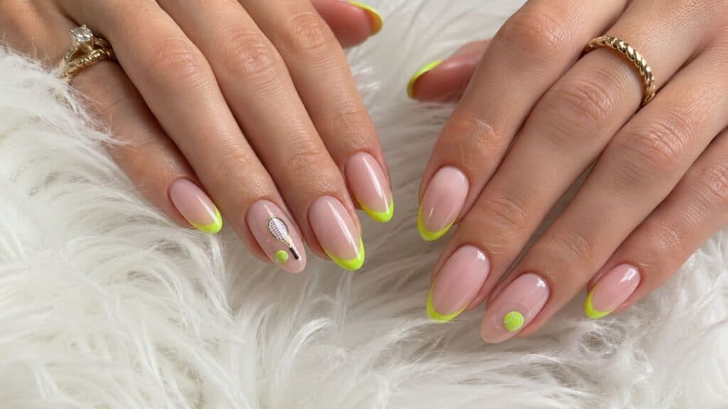 Hands with almond-shaped nails featuring nude polish, neon yellow tips, and minimalist nail art accents, resting on a white fluffy surface.