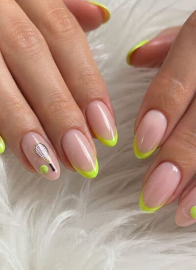 Hands with almond-shaped nails featuring nude polish, neon yellow tips, and minimalist nail art accents, resting on a white fluffy surface.